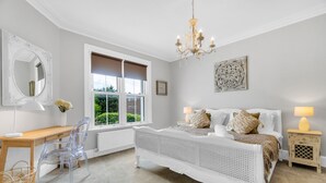 Une chambre avec un grand lit, un bureau en bois, une chaise, une fenêtre avec des stores, un lustre et une petite table de chevet avec une lampe.
