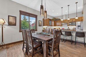 A modern kitchen with wooden cabinets, a dining table with chairs, pendant lighting, and a view of the outdoors through a window.