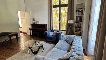 Een woonkamer met een grijze bank, een glazen salontafel, een boekenkast en een raam met gordijnen.