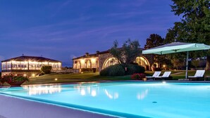 A poolside view of a property with a large pool, lounge chairs, and a well-lit building with arched windows.