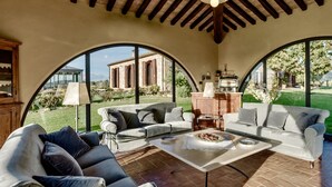 A spacious living area with two sofas, a coffee table, and a kitchen area with a stove. Large arched windows provide views of a garden and a building outside.
