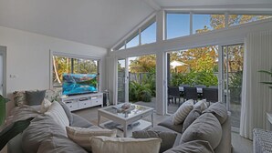A modern living room with a large flat-screen TV, a sofa, a coffee table, and a sliding glass door leading to an outdoor patio with chairs and plants.