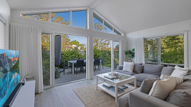 A spacious living room with a large sofa, a coffee table, and a TV. There is a sliding glass door leading to an outdoor patio area with a table and chairs.