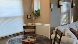 Un coin repas avec une table ronde, des chaises et un lustre. On y trouve une fenêtre avec des stores, une plante et une étagère fixée au mur, sur laquelle sont posées des bougies.