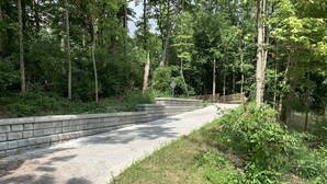 Un chemin en béton dans une zone boisée, bordé d’un mur de pierres et d’une clôture en bois.