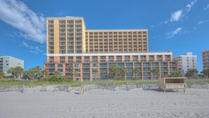 Ein Hotel mit mehreren Etagen, einem Sandstrand davor und einem strahlend blauen Himmel.