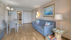 Ein Wohnzimmer mit einem blauen Sofa, einem Glastisch, einem Esstisch mit Stühlen und einer Kristallleuchter.
