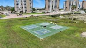 A tennis court surrounded by grass, with a parking lot and buildings in the background. - Bonita Beach and Tennis Unit 2808 (Bonita Springs)