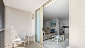 Un balcón con una silla y una mesa blancos, y un ventanal corredero que da a una sala de estar con un sofá y una cocina al fondo.