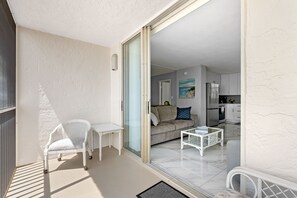 Ein Balkon mit weißem Stuhl und Tisch, eine Schiebetür zu einem Wohnbereich mit Sofa und Küche im Hintergrund.