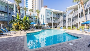 Piscina con sillones y sombrillas, rodeada de edificios de varios pisos con balcones.