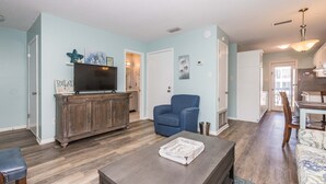Una sala de estar con televisión, un sillón azul, un aparador de madera y un comedor con mesa y sillas.