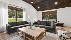 Una sala moderna con techo de madera, un ventanal grande, un sofá gris, una mesa de centro de madera y una zona de comedor con mesa y sillas.