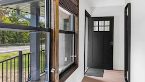 Un pasillo con una puerta negra, una ventana con marco de madera y vista a una calle y árboles.