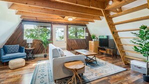 Une chambre à coucher confortable, aménagée en mezzanine, avec un plafond en pente, des poutres en bois, un canapé, une télévision et une grande fenêtre donnant sur les arbres.