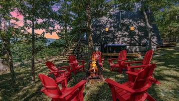 Ein Lagerfeuer mit roten Adirondack-Stühlen darum herum, eine Holzterrasse und eine Treppe zu einem Gebäude.