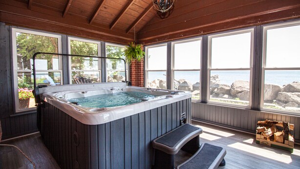 A hot tub in a wooden room with large windows overlooking a rocky shoreline.