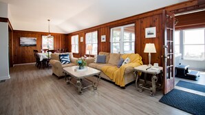 A spacious living room with wooden paneled walls, a ceiling fan, a dining area with a table and chairs, and a cozy seating area with a sofa and armchairs.