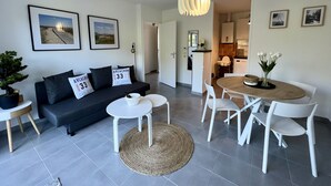 Una sala de estar moderna con un sofá, una mesa de centro y una mesa de comedor. Hay cuadros enmarcados en la pared, una planta en maceta y una lámpara colgante.