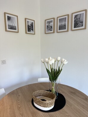 Pyöreä puinen pöytä, jonka päällä on punottu kori ja valkoisia tulppaanikukkia sisältävä maljakko. Pöydän yläpuolella seinällä on neljä kehystettyä mustavalkokuvaa.
