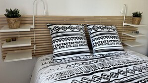 Una cama con colcha estampada, dos almohadas con diseños geométricos, una mesita de noche con una cesta tejida y un estante de pared con una planta.