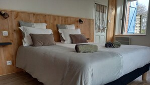 A neatly made bed with a wooden headboard, a window with curtains, and a view of the outside. - Single-storey house 100 m from the strikes (Courtils)