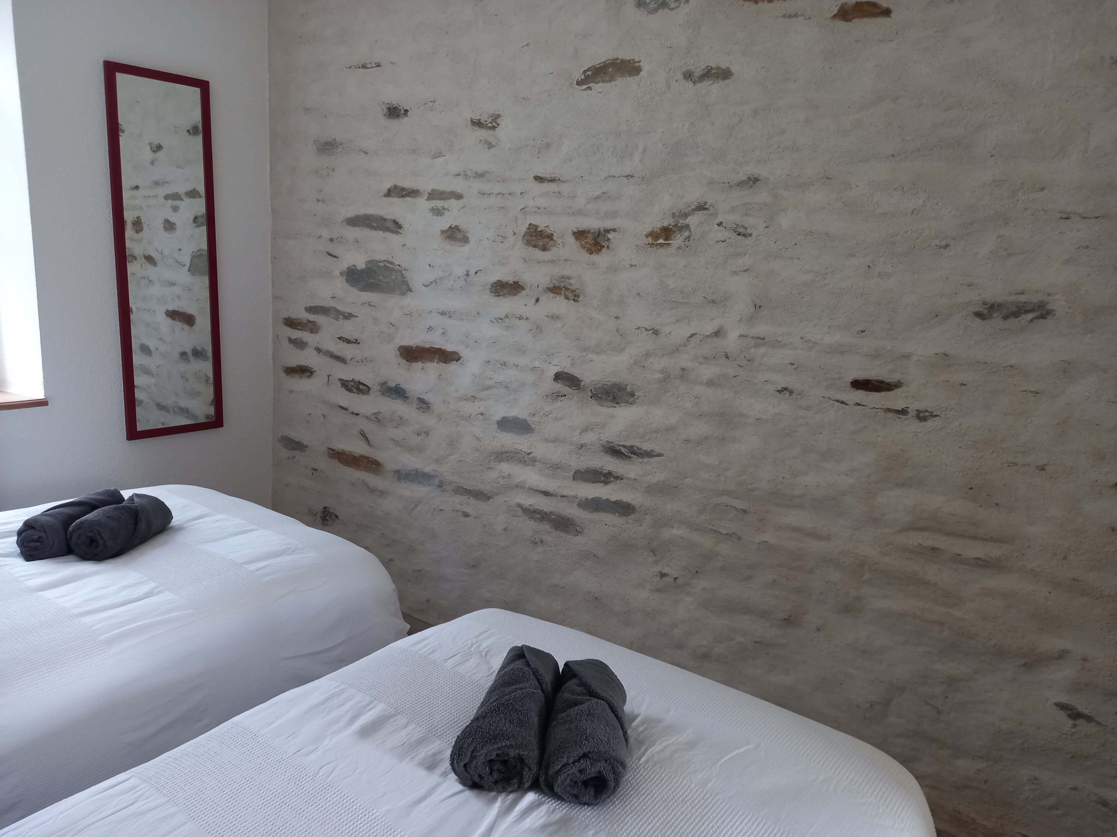 Two beds with white linens and rolled towels, a red-framed mirror, and a textured stone wall.