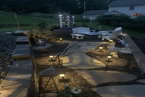 Een terras buiten met stenen vloer, witte tuinmeubels en decoratieve verlichting.