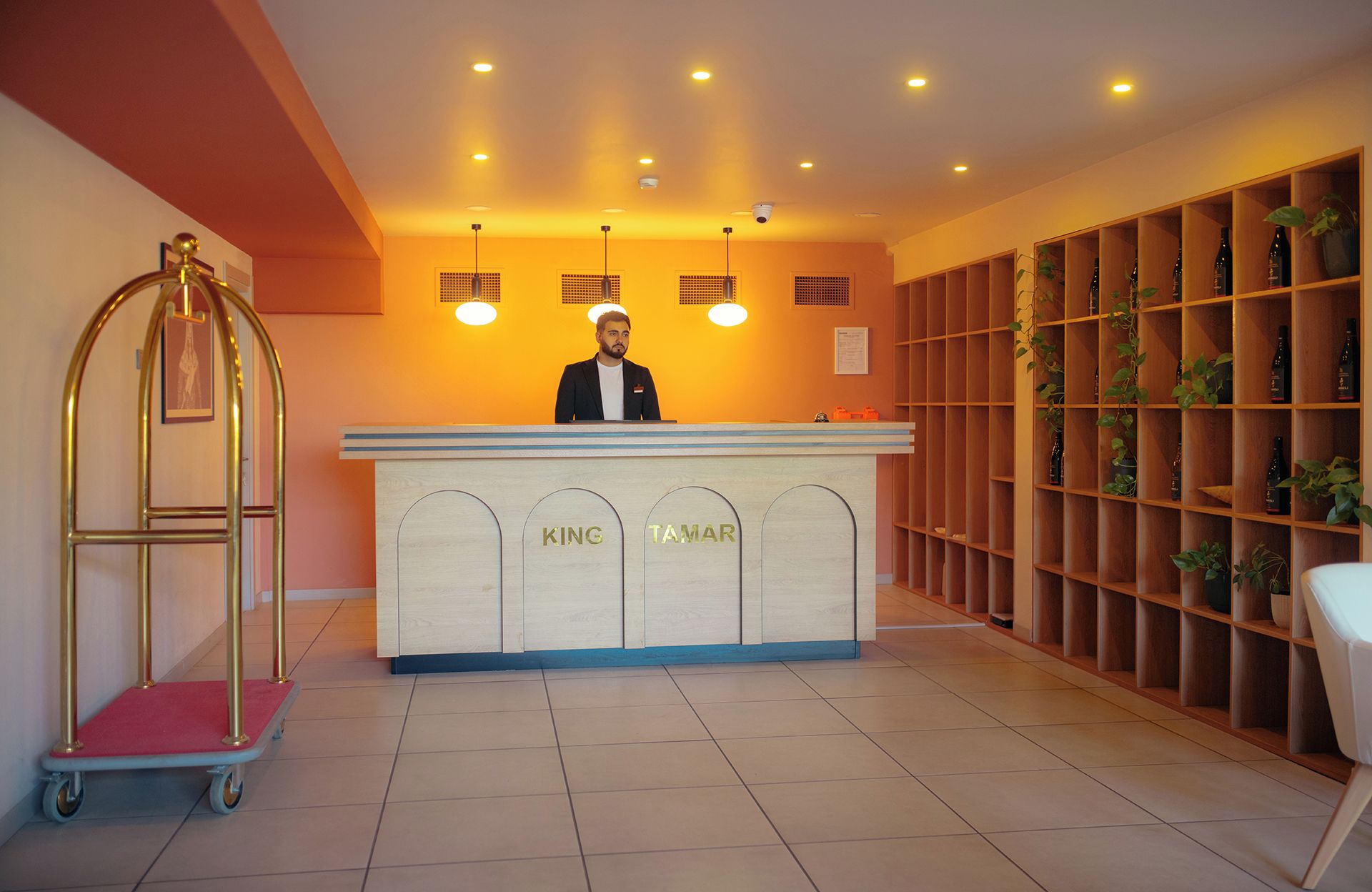 A reception desk with 'KING' and 'TAMAR' signage, a cart, and a shelving unit with bottles and plants.