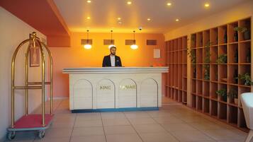A reception desk with 'KING' and 'TAMAR' signage, a cart, and a shelving unit with bottles and plants.
