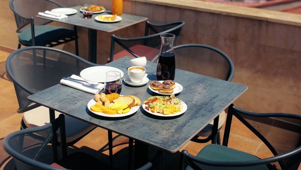 Een ontbijttafel met etenswaren, een fles donkere vloeistof en een glas sinaasappelsap, met uitzicht op een stadsgezicht met gebouwen en groen.