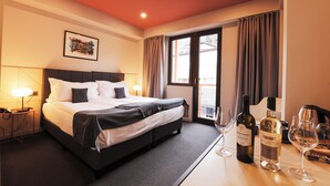 Een hotelkamer met een groot bed, een bureau met wijnglazen en flessen, een lamp en een raam met gordijnen.