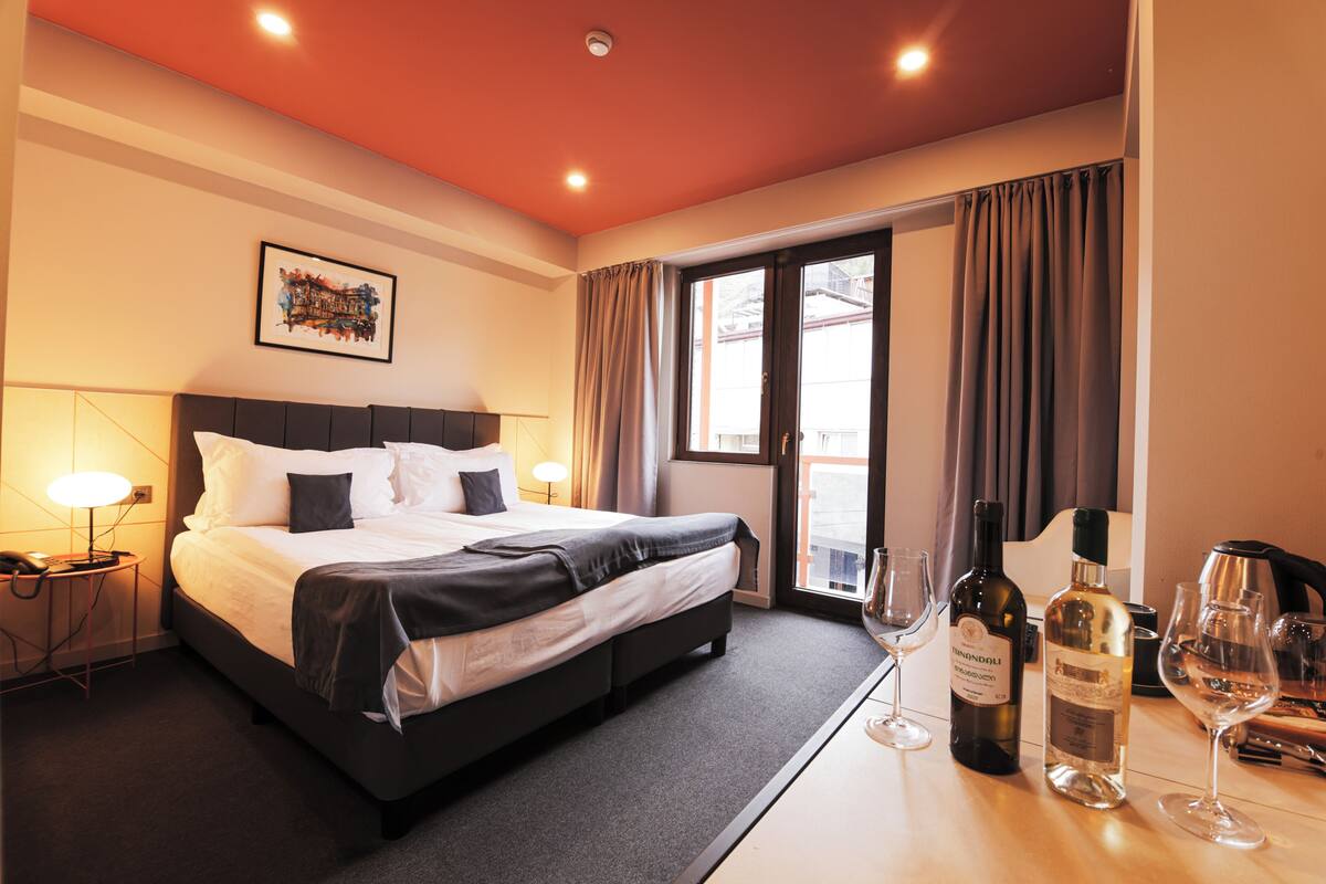 A hotel room with a large bed, a desk with wine glasses and bottles, a lamp, and a window with curtains.