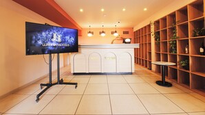 Een receptiezone met een flatscreen-tv waarop de branding 'LUXURIA HOTELS' te zien is, een receptiebalie met de aanduiding 'KING' en 'TAMAR', en een wand met houten planken met planten en flessen.