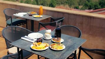 Une table dressée avec des aliments pour le petit-déjeuner, une bouteille contenant un liquide sombre et un verre de jus d’orange, donnant sur un paysage urbain avec des bâtiments et de la verdure.