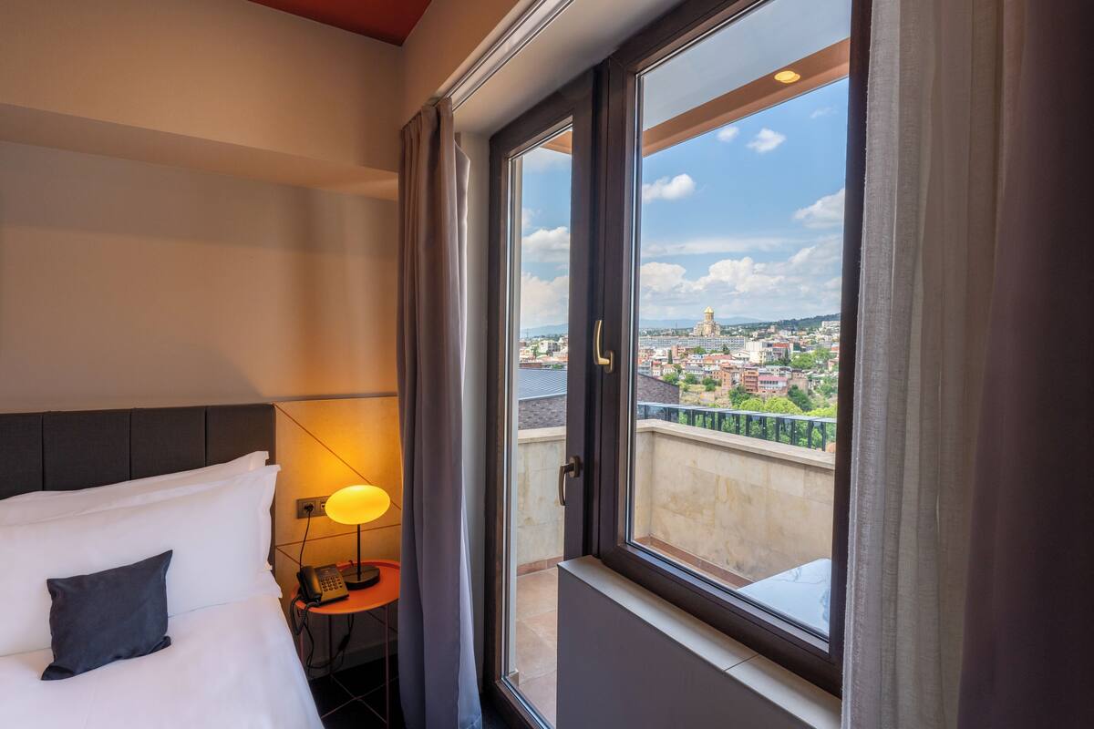 A hotel room with a bed, a bedside lamp, a phone, and a window with a city view.