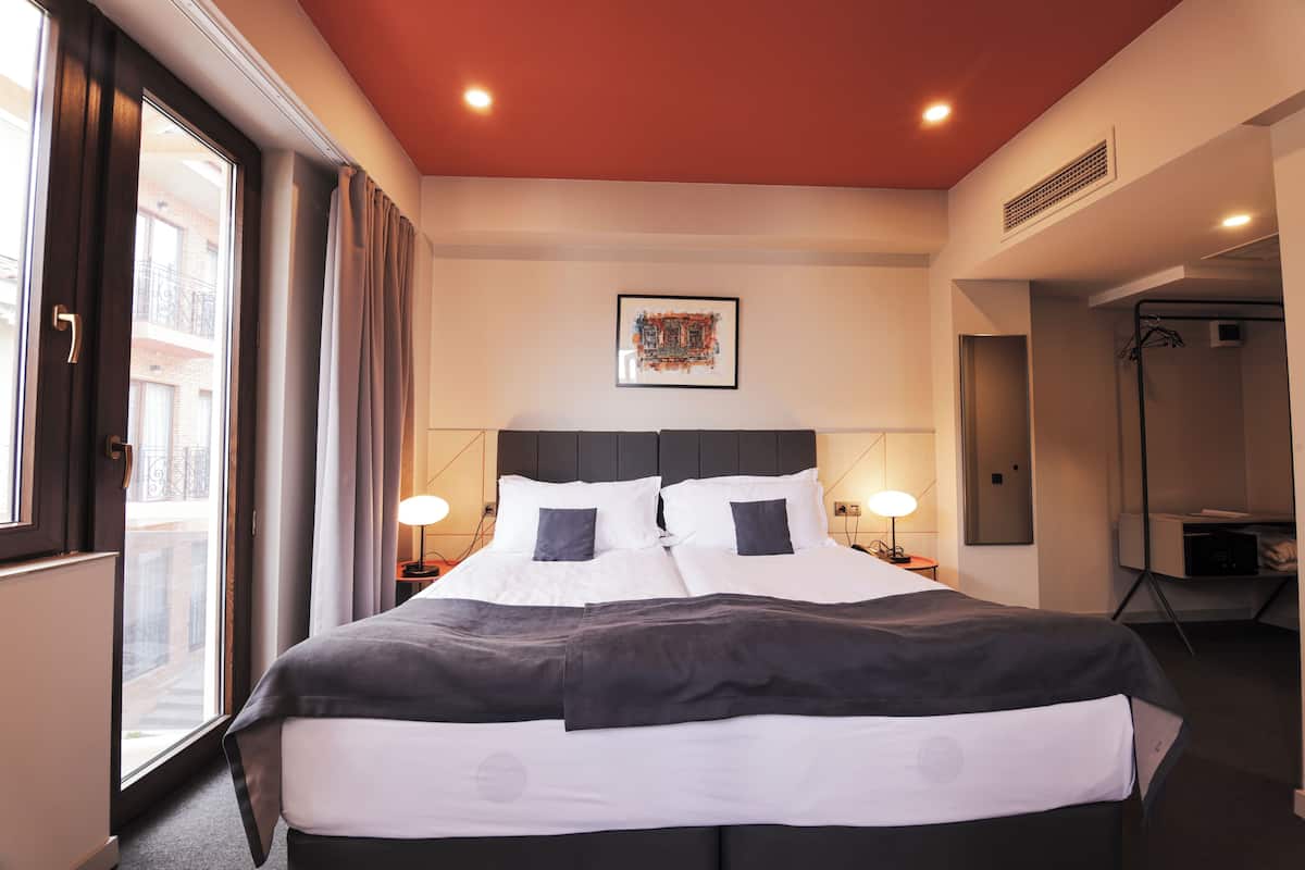 A hotel room with a large bed, two bedside tables, a window with curtains, a ceiling light, a framed picture on the wall, and a door leading to a bathroom.