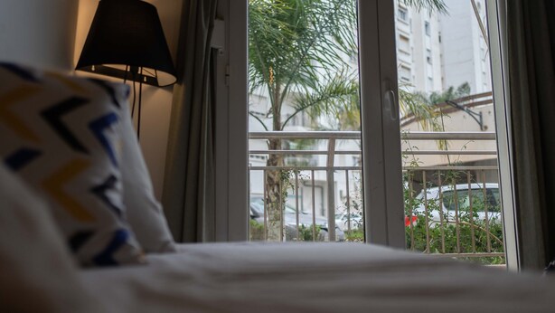 A hotel room with a bed, a window with a view of a palm tree and a building, and a lamp on the wall. - Hotel comfort in a prestigious residence (Chéraga)