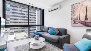 Ein modernes Wohnzimmer mit einem grauen Sofa, einem runden Couchtisch und einem großen Fenster mit Blick auf eine Stadtlandschaft.