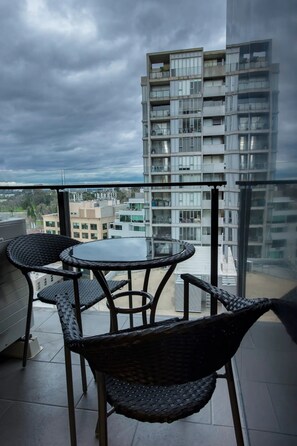 Ein Balkon mit Korbstühlen und einem Glastisch, mit Blick auf ein modernes Gebäude und die Stadtlandschaft.