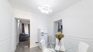A modern, minimalist dining area with a glass table and white chairs, a white refrigerator, and a sofa in the background. - iRoome Rome (Roma)