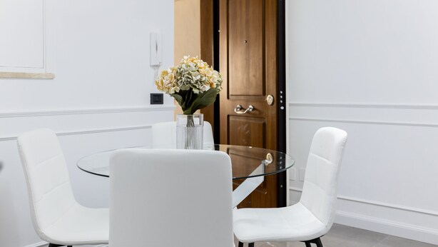 A modern dining area with a glass table and white chairs, a vase with flowers, and a wooden door in the background. - iRoome Rome (Roma)