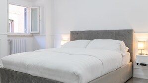 A modern bedroom with a grey upholstered bed, white bedding, and bedside lamps. - iRoome Rome (Roma)