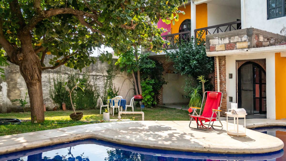 Una piscina amb gandules, una taula i un arbre a un pati.