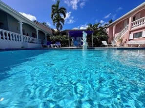 A clear blue pool with lounge chairs and a statue in the center, surrounded by palm trees and a building with a balcony. - Detached yellow studio in front of the pool 150m from the beach! (saint martin)