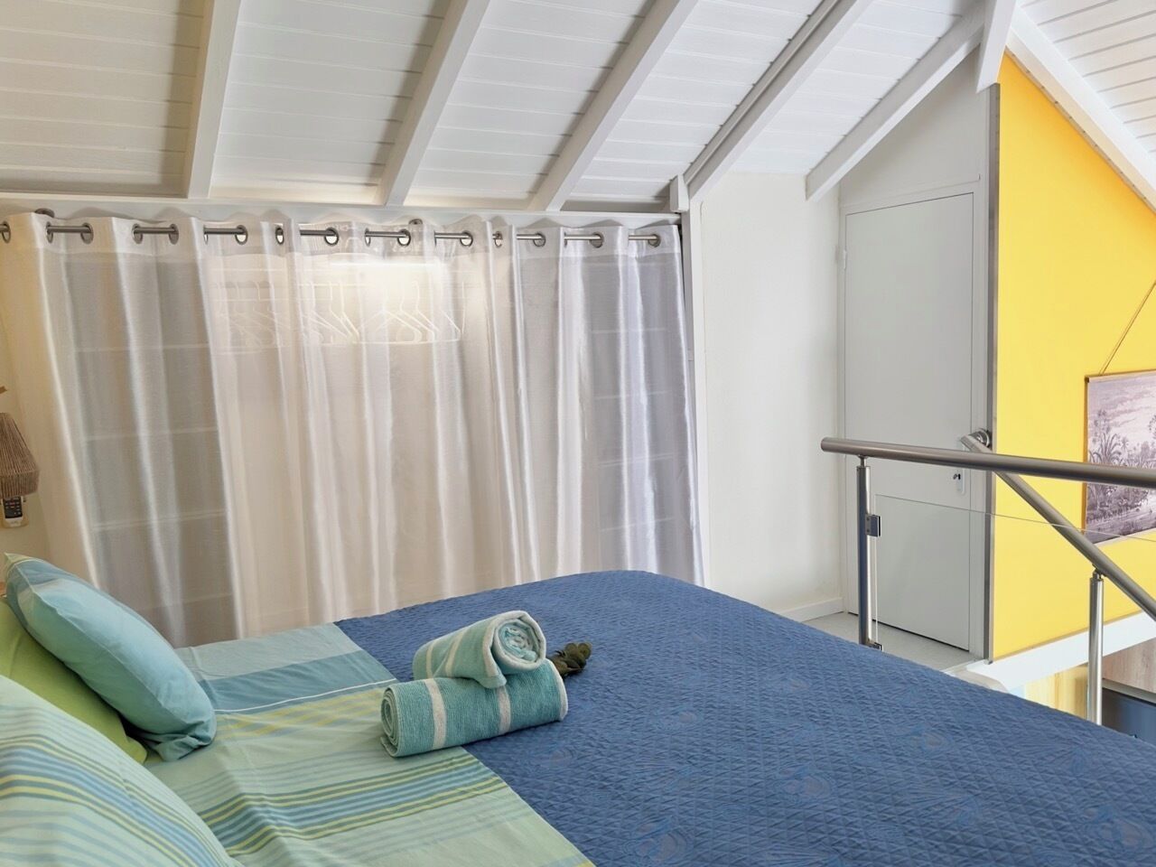 Een slaapkamer met een bed, kussens, een opgerolde handdoek, een raam met gordijnen, een deur en een gele muur.