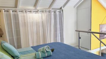 Een slaapkamer met een bed, kussens, een opgerolde handdoek, een raam met gordijnen, een deur en een gele muur.