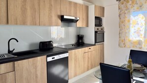 A modern kitchen with wooden cabinets, a black countertop, a dishwasher, a coffee maker, and a dining area with a glass table and chairs. - Detached yellow studio in front of the pool 150m from the beach! (saint martin)