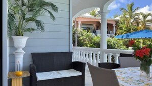 A covered outdoor seating area with a wicker sofa, a table with a floral tablecloth, and potted plants. - Detached yellow studio in front of the pool 150m from the beach! (saint martin)