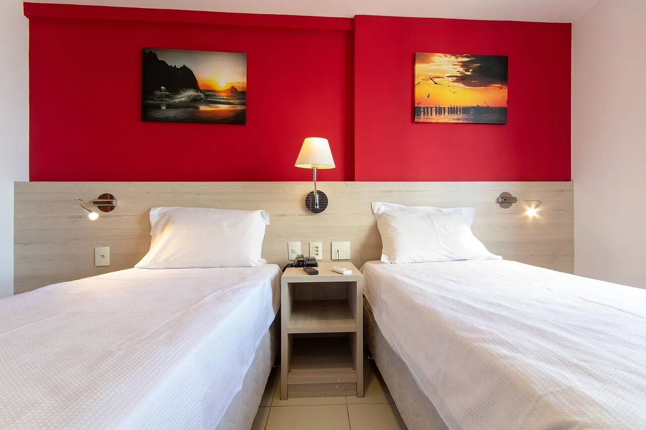 A hotel room with two beds, a red accent wall, and two sunset paintings.